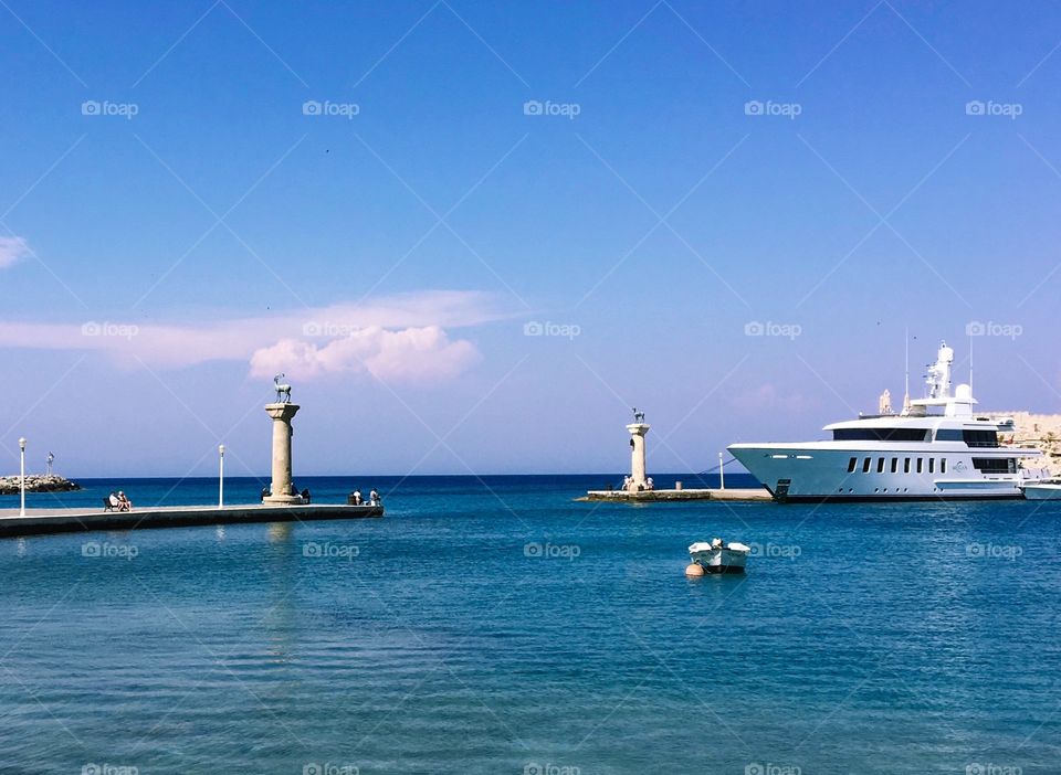 There was a boat and a yacht anchored on the harbor, the water was calm and beautiful in blue, purple cloudy on blue sky, there are 2 deer statues on 2 pillars. 