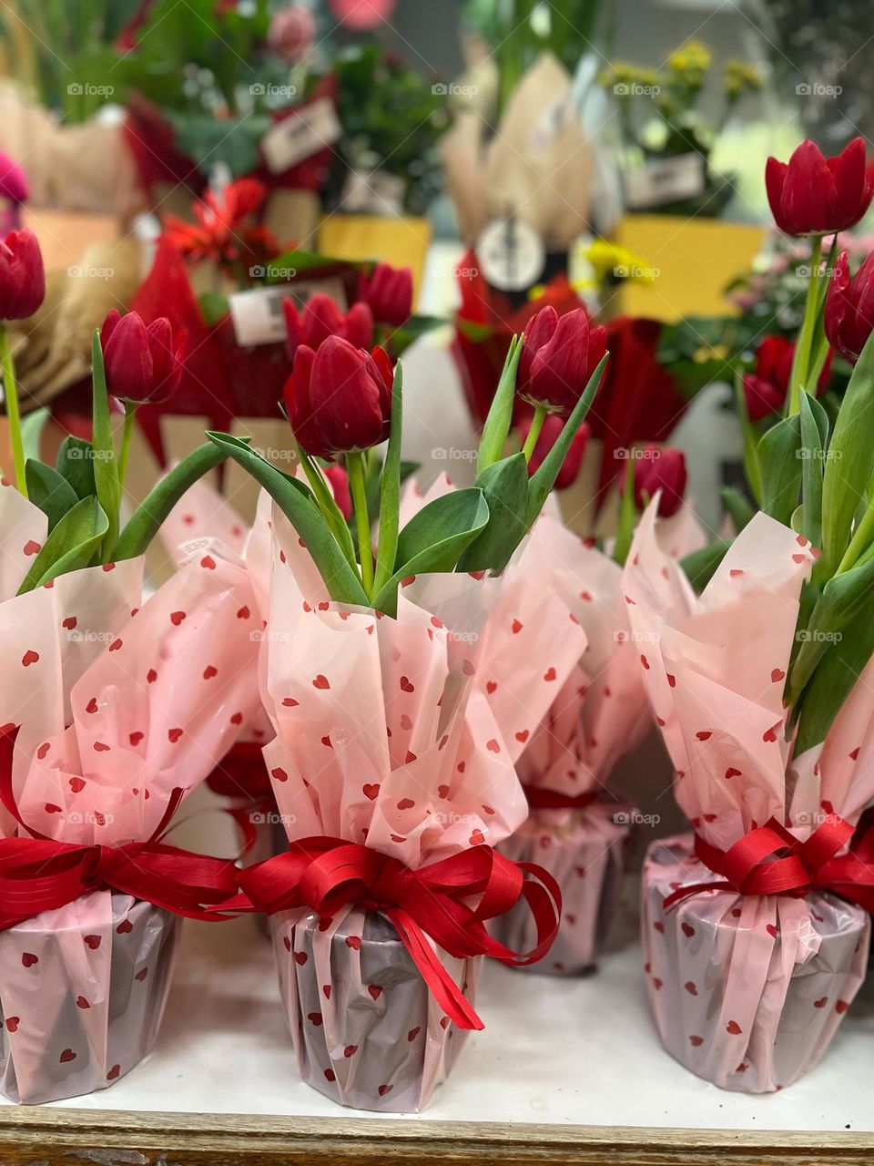 Vibrant red tulips elegantly wrapped in pink paper with a heart pattern, attached with red ribbons, attractively arranged as romantic gifts or Valentine's Day gifts, surrounded by other floral arrangements.