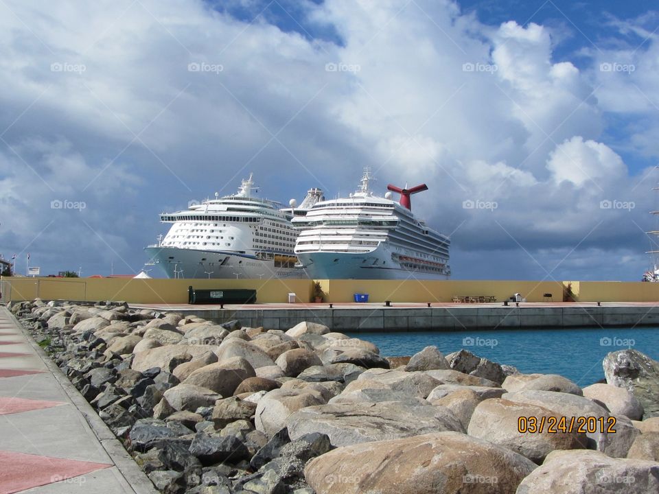 Cruise ships docked side by side in the distance, rock jetty in foreground of Caribbean water.