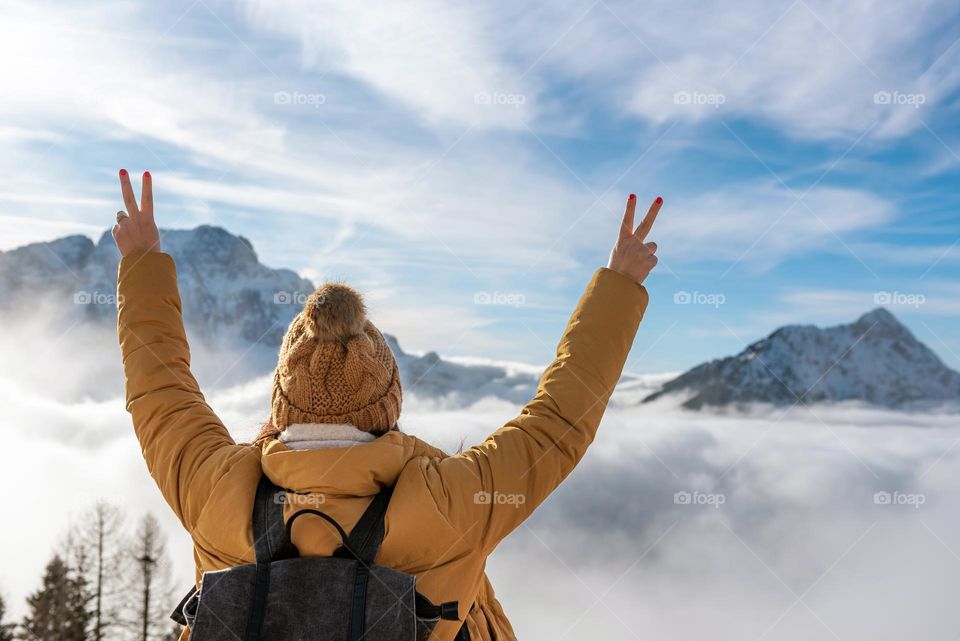 Rear view of young woman with raised arms wearing a backpack and winter clothes admiring mountain peaks and clouds on a sunny day in winter