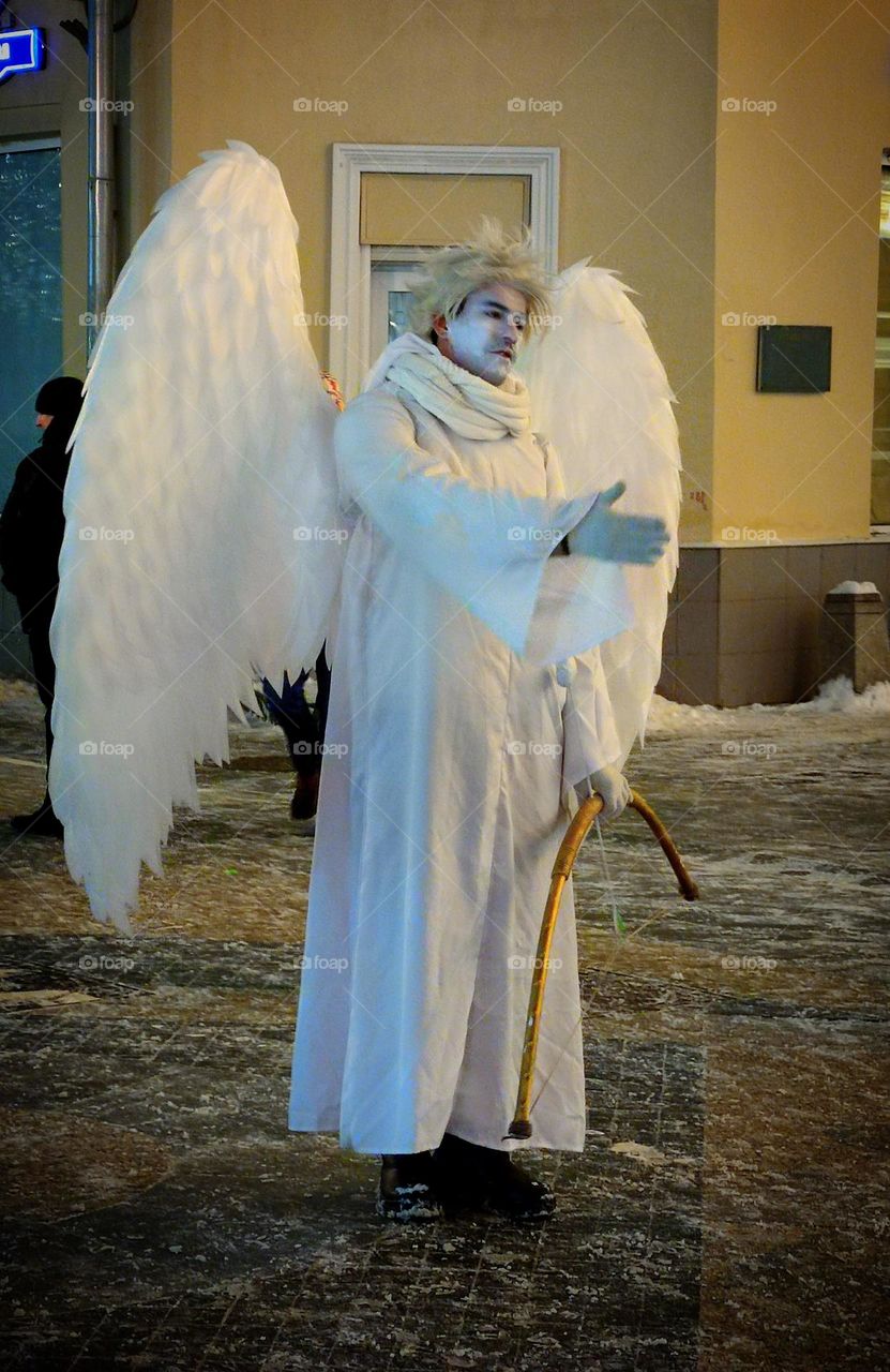 Sadness.  A man dressed as a white angel is standing on the street.  The angel holds out one hand to people.  The face and posture of the angel shows the emotion of sadness