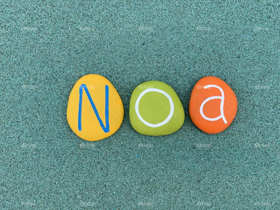 Noa, feminine given name composed with multi colored stones over green sand 