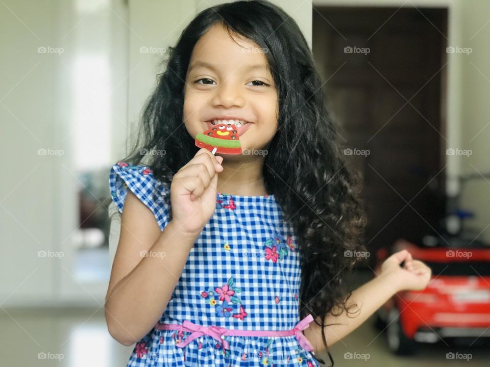 Girl enjoying a lot by eating candy lollipop and she smiles very gracefully and she wears blue colour frock and leaves her hair.looks 😎