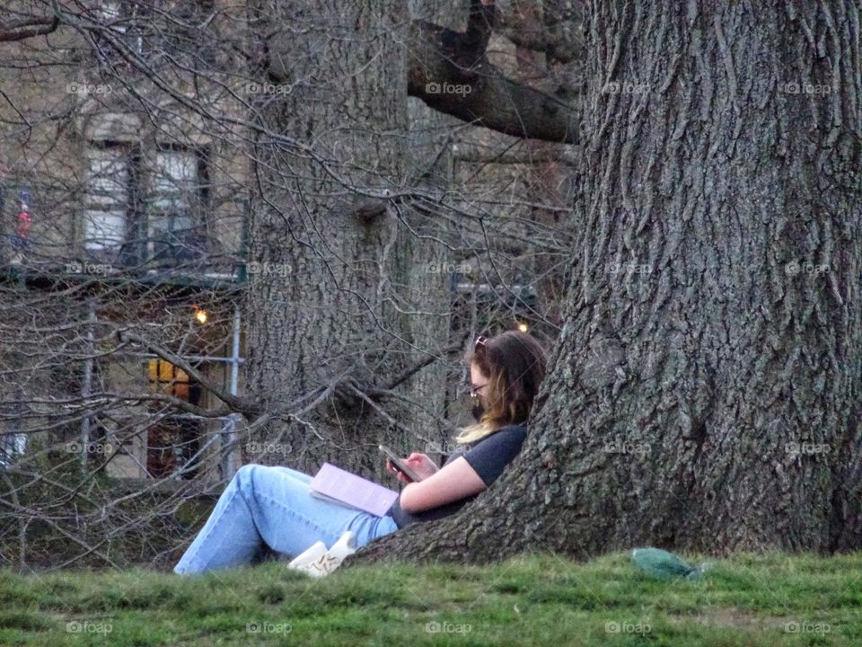 A woman leaning on the tree trunk holding a smartphone and a book on her lap.