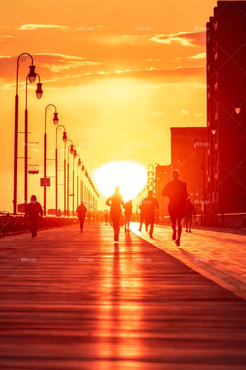 Silhouettes of people jogging down a boardwalk during a beautiful orange sunset, as the sun lines up perfectly down the path. Telephoto focal length compression. 