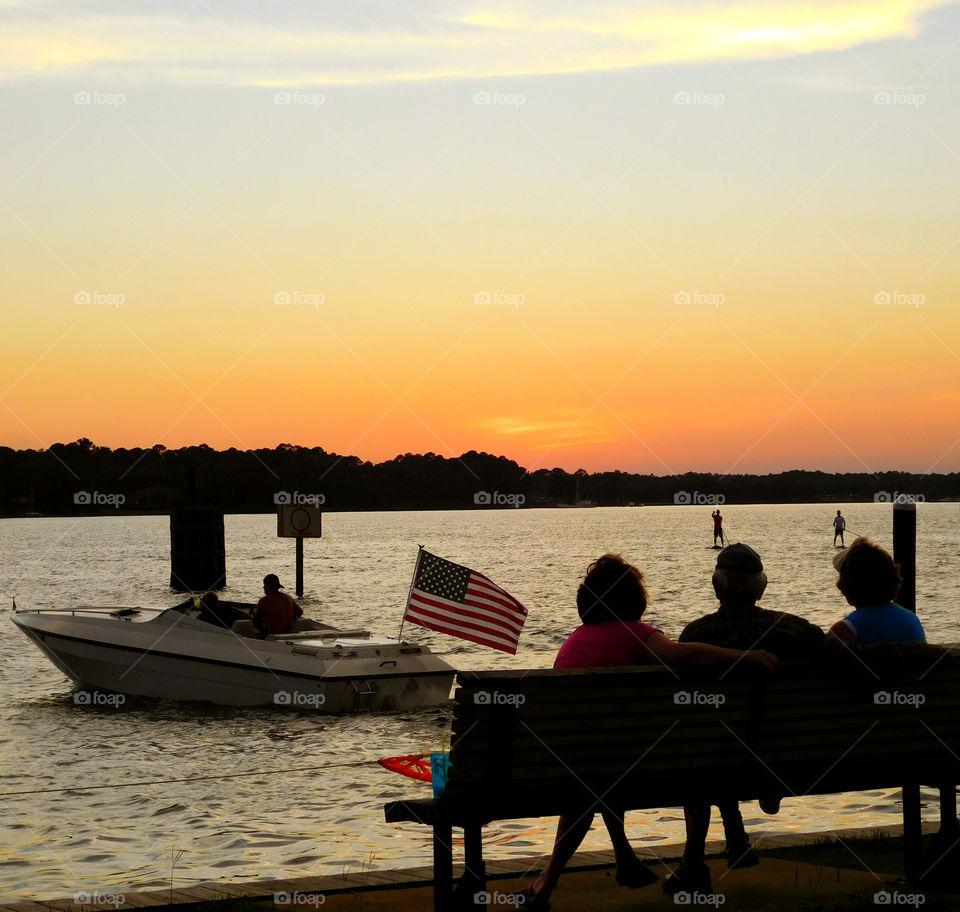 Boater leaves the dock with the American flag flying as people sit on the park bench admiring the sunset! Two men paddle board toward the dock after a day on the bayou!