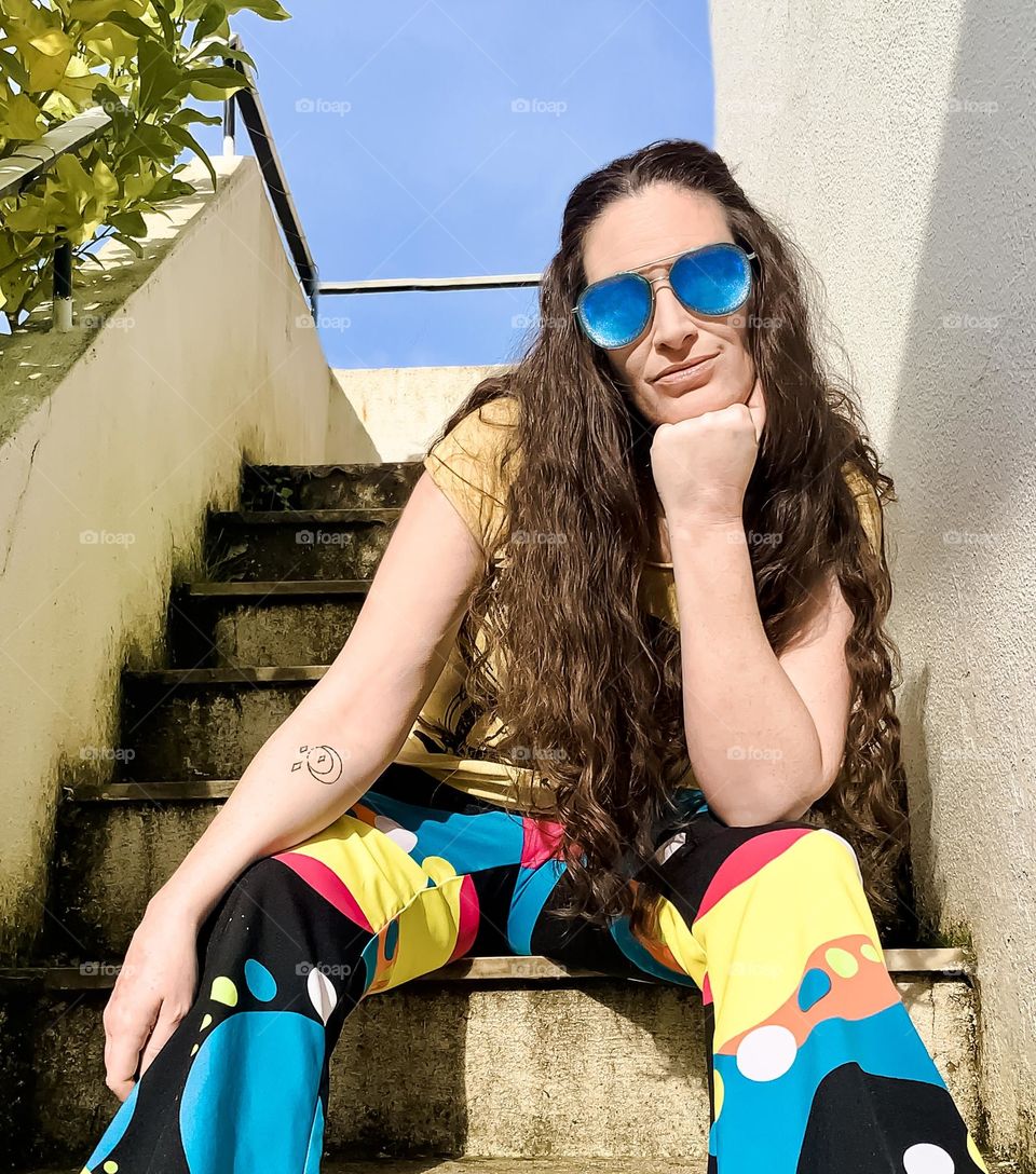 Woman with long hair, wearing psychedelic flares and blue sunglasses sits on steps in the sunshine 