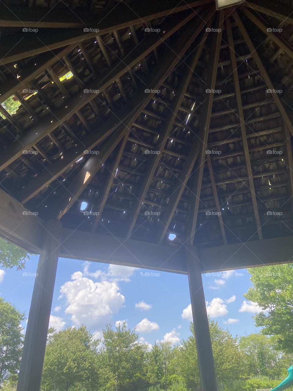 View of bright blue sky, puffy white clouds and green trees from the shady shelter of a gazebo on a hot summer day. Taken in a park in Bay Head, NJ.