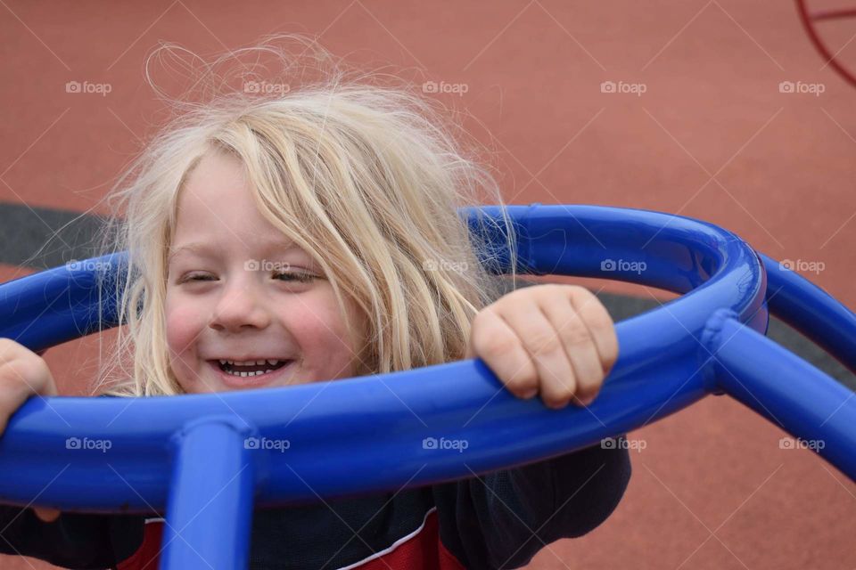 Toddler boy playing in a bright blue spinning toy at the playground. The little boy has long blonde hair.