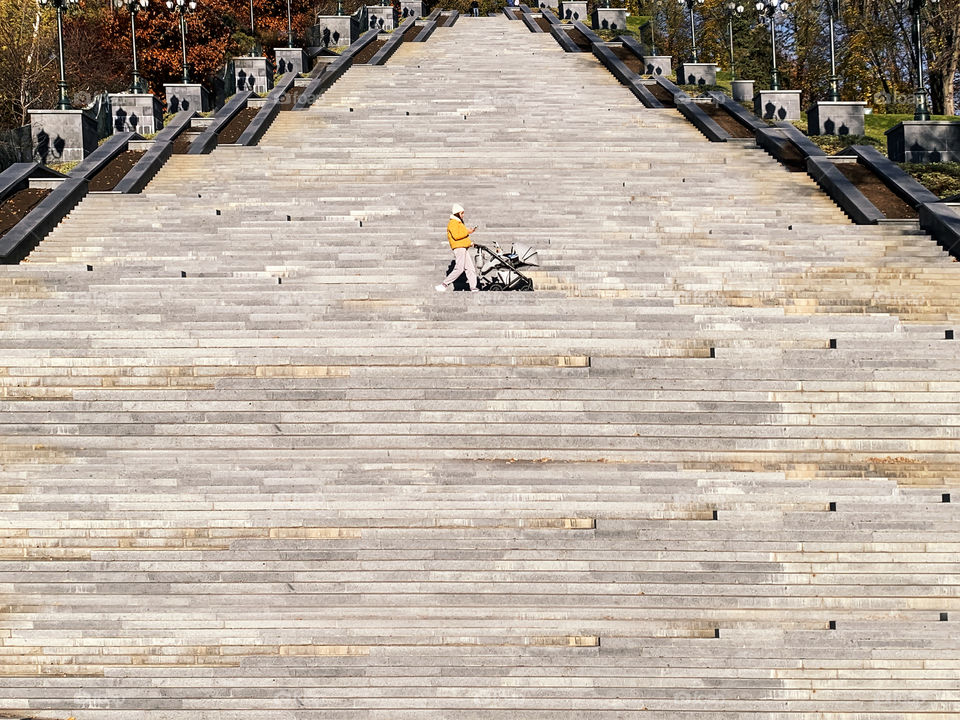 Tiny figure of a young woman in yellow jacket with baby carriage walking by huge staircase 