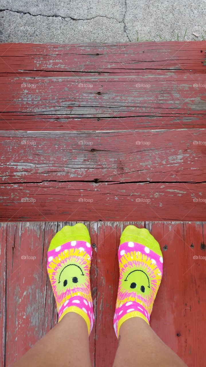 Standing at the top of wood stairs. Looking down at my feet with happy socks on. At the bottom of the stairs is concrete walkway.