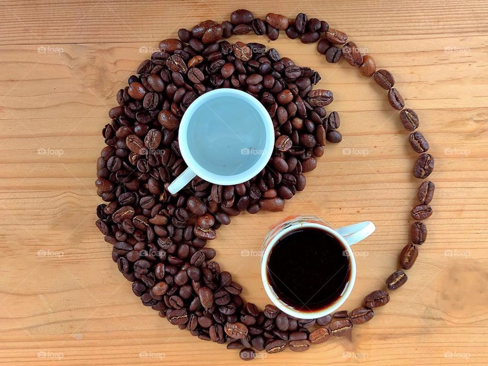 Philosophy. On a wooden background lie coffee beans in the shape of a yin-yang symbol. The symbol is complemented by a white cup on coffee beans and a cup of black coffee on a wooden surface.