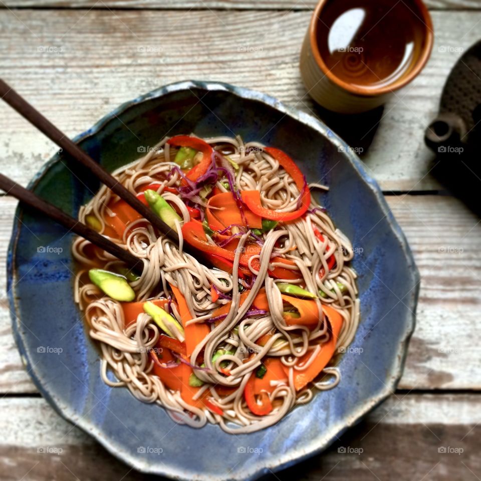 Noodles with sliced carrots and red bell peppers in broth on a rustic wood table with a cup of tea.