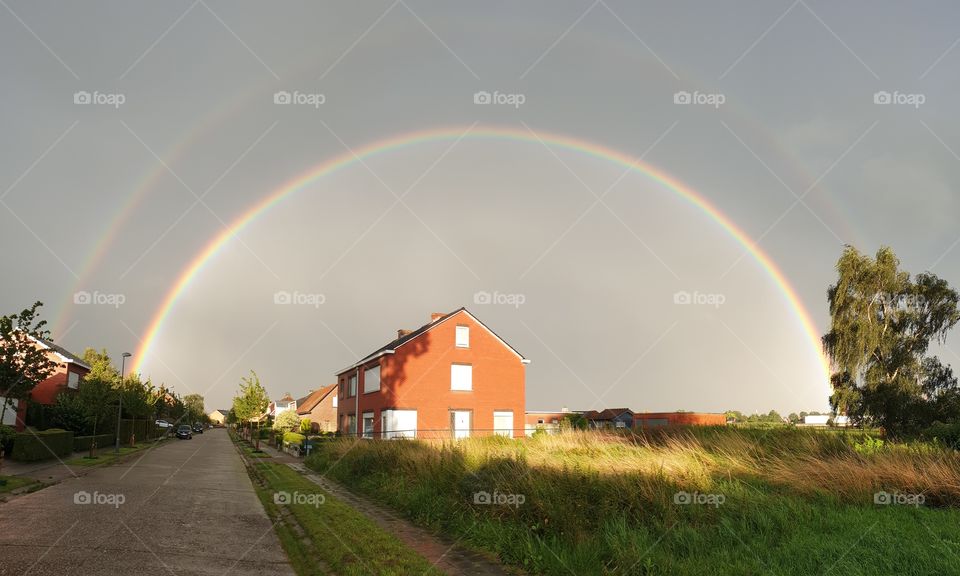 a portrait of a double rainbow in the sky. the rainbows are above a field house and street. the lowest one is very clear the other one is a bit vague