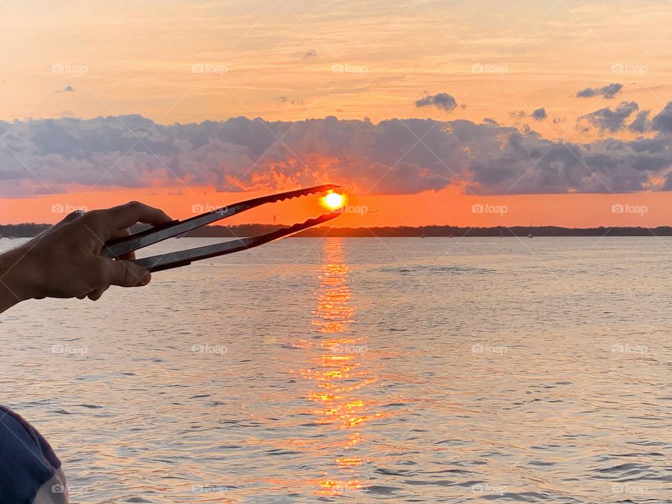 Sun From Sunset Seems To Be Grabbed By The BBQ Tongs  On Top Of An Island In The Horizon At The Seashore On A Tropical Environment.