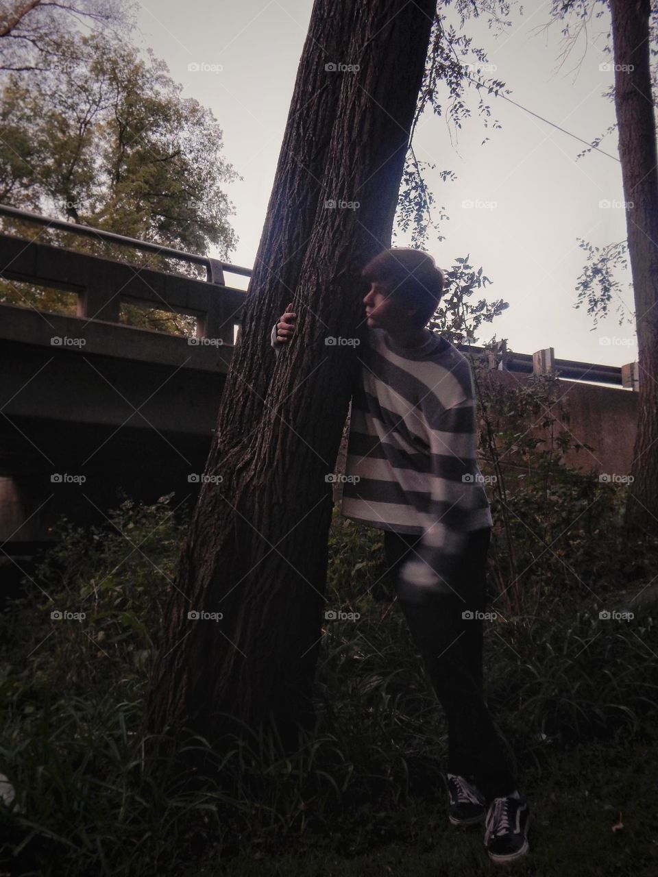 Young man wearing long sleeved black and white striped shirt and black jeans leaning against a tree near a bridge that has green vines and weeds growing around it under an overcast gray sky in Michigan