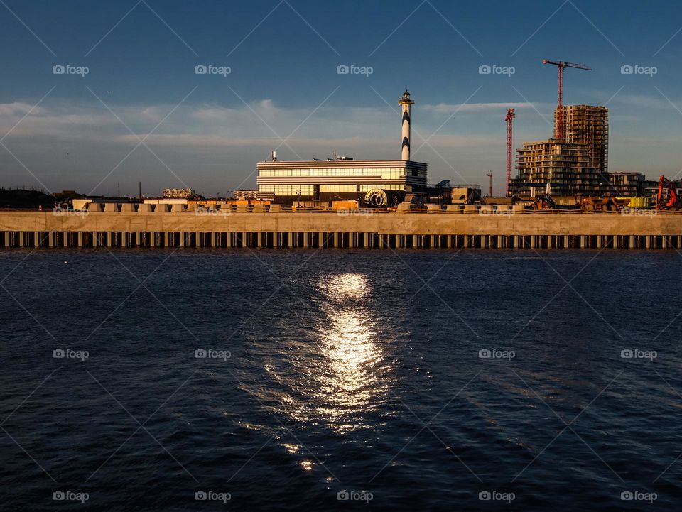 A beautiful view of the evening sunset landscape of the northern sea with reflection and glare of the sun on the water and a lighthouse, buildings and a crane in the distance, side view close-up.