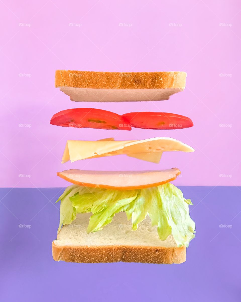 Levitation sandwich: bread, lettuce, ham, cheese, tomato. Pink and violet background.