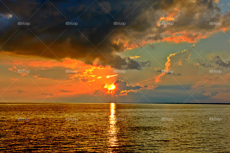 Painted Sky. The sunset radiates behind a thick, heavy storm cloud.  The sun rays reflect off the traveling clouds and the waving bay