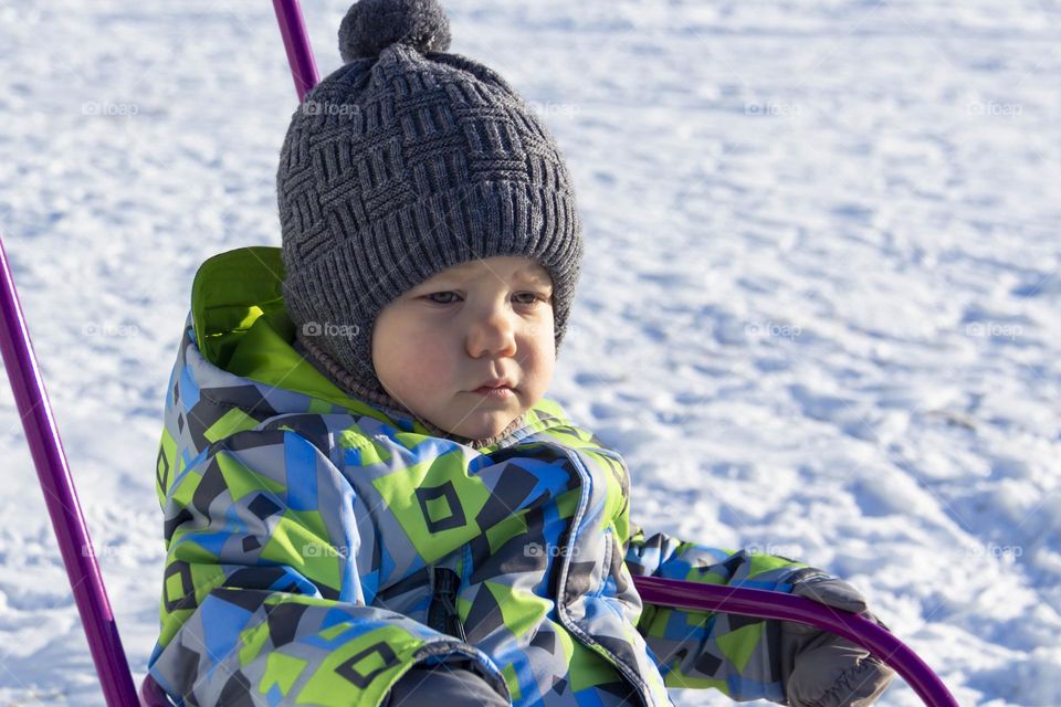 A child with a serious expression on his face in winter clothes jackets, pants, hat and boots in winter on white snow on the street and in the park in nature sledding and playing winter fun.
