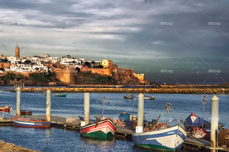 Fishing boats at the pier on the river Bu Reger (Rabat, Morocco) against the background of the old fortress and flocks of gulls circling over the mouth of the river under a pre-storm cloudy sky on a clear spring morning