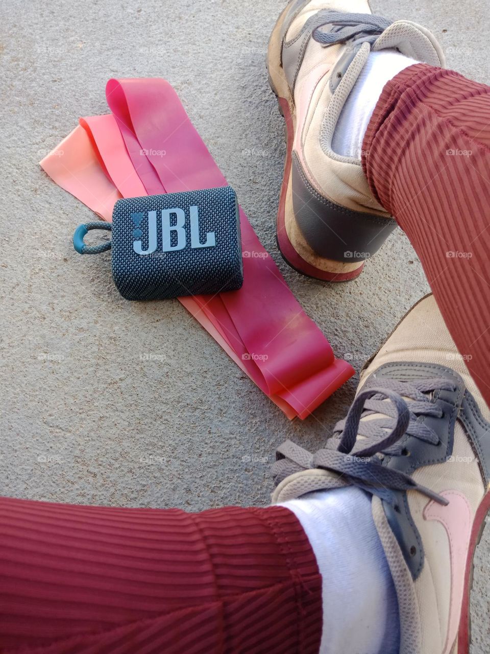 Pink exercise bands,  JBL go box, Nike shoes grey and pink,  partial legs,  grey floor