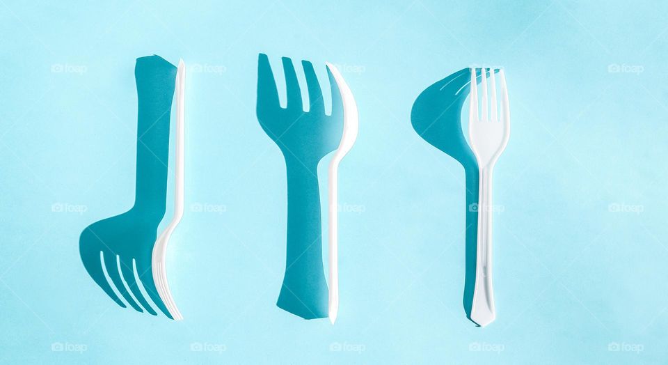 Three white plastic forks with hard shadows from the sun lie in the center on a blue background, flat lay close-up.Cutlery and ecology concept.