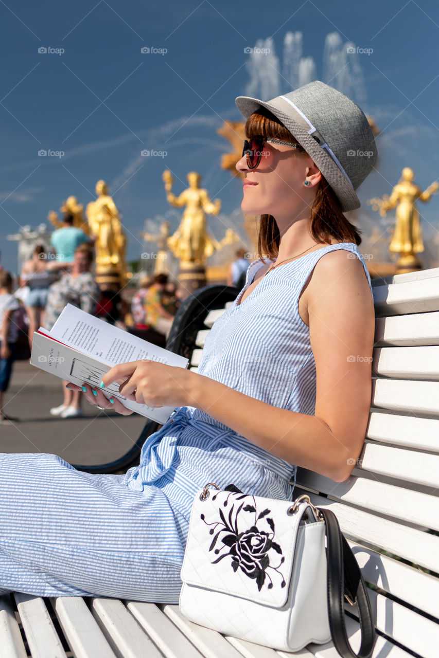 A girl sits on a bench with a book in the VDNH park near the fountain of friendship of nations. Moscow city.