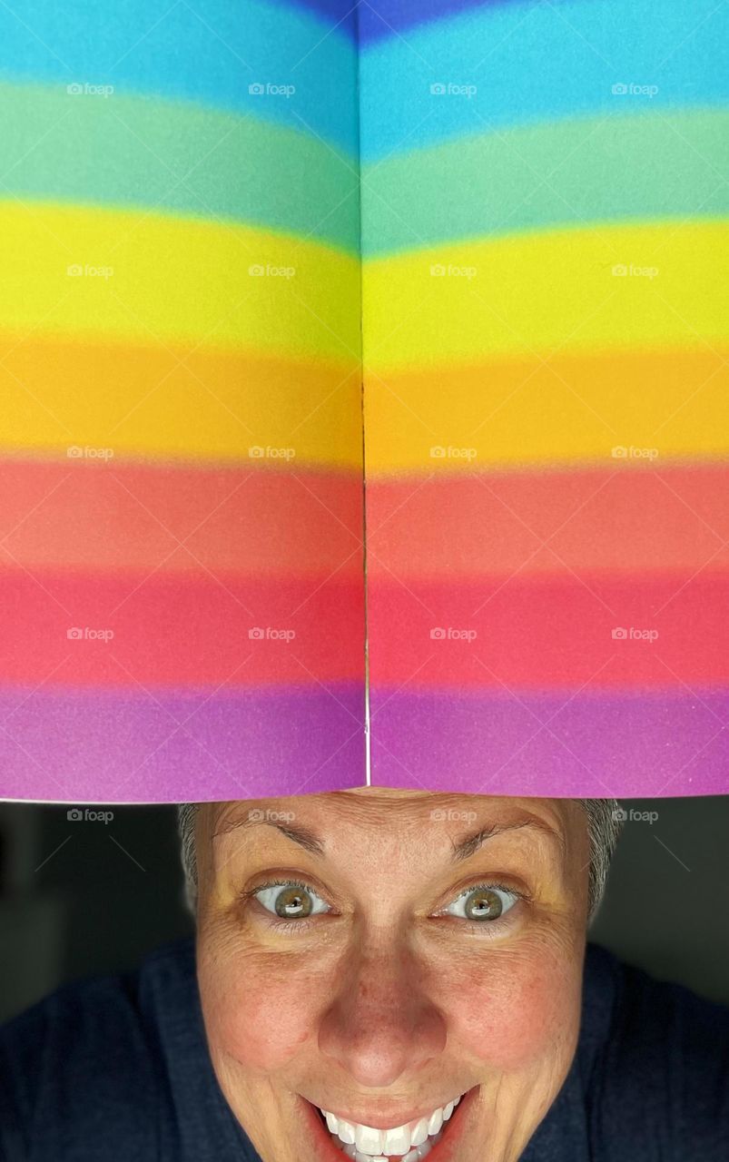 Woman with pride colored book, woman peeks out from behind a rainbow, woman coming out from behind a rainbow, Pride colors in a children’s book, woman comes out in Pride month