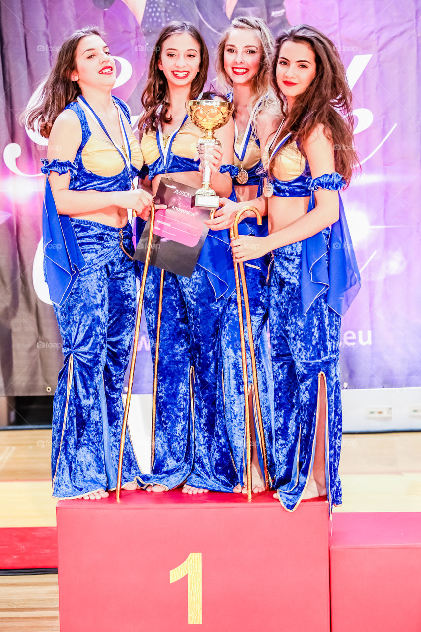 Beautiful Young Happy Girls Winning Golden Trophy Award At Number One Place In Podium
