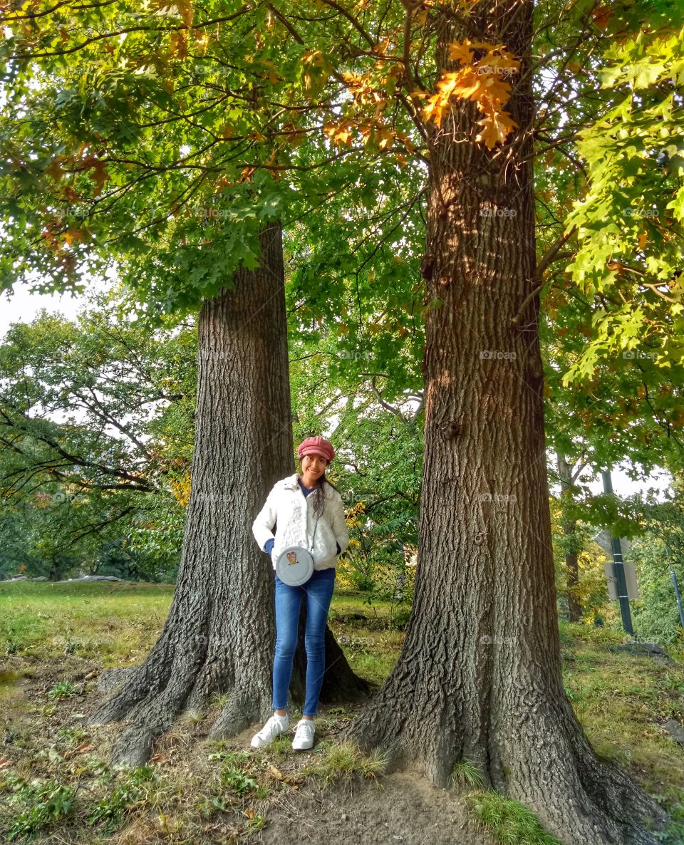 A woman wearing a red hat, white sweater. Hands on her pocket, standing with two big trees background. 