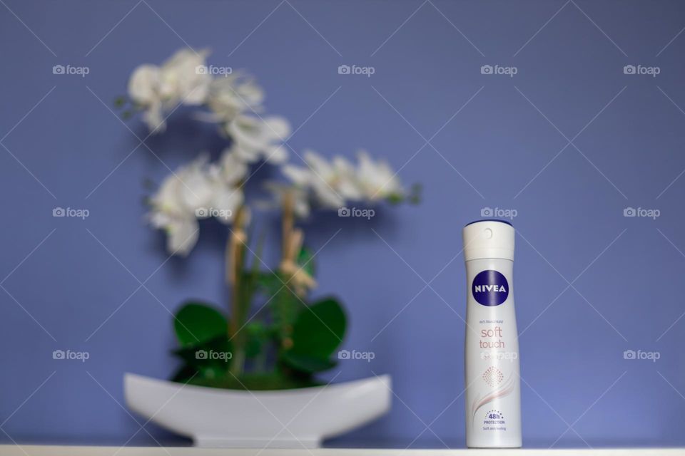 One bottle of Nivea brand anti transpirant stands on a white chest of drawers with a potted orchid flower blurred behind against a blue-lilac wall, close-up view from below.