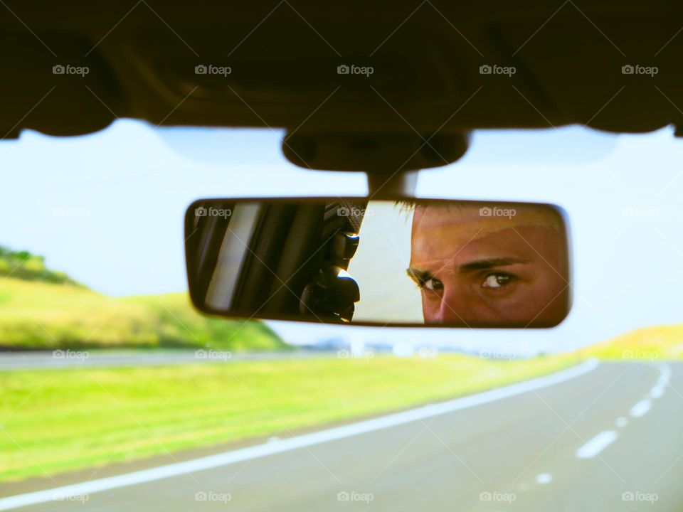 Man with green eyes driving, looking at the rear mirror, with the road sprawling ahead. A road trip photo.
