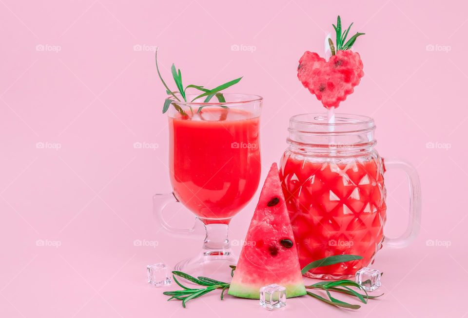 Fresh watermelon drink in glasses with a piece of triangle and heart shaped watermelon, rosemary sprigs, straws and ice cubes on a pink background, close-up side view.