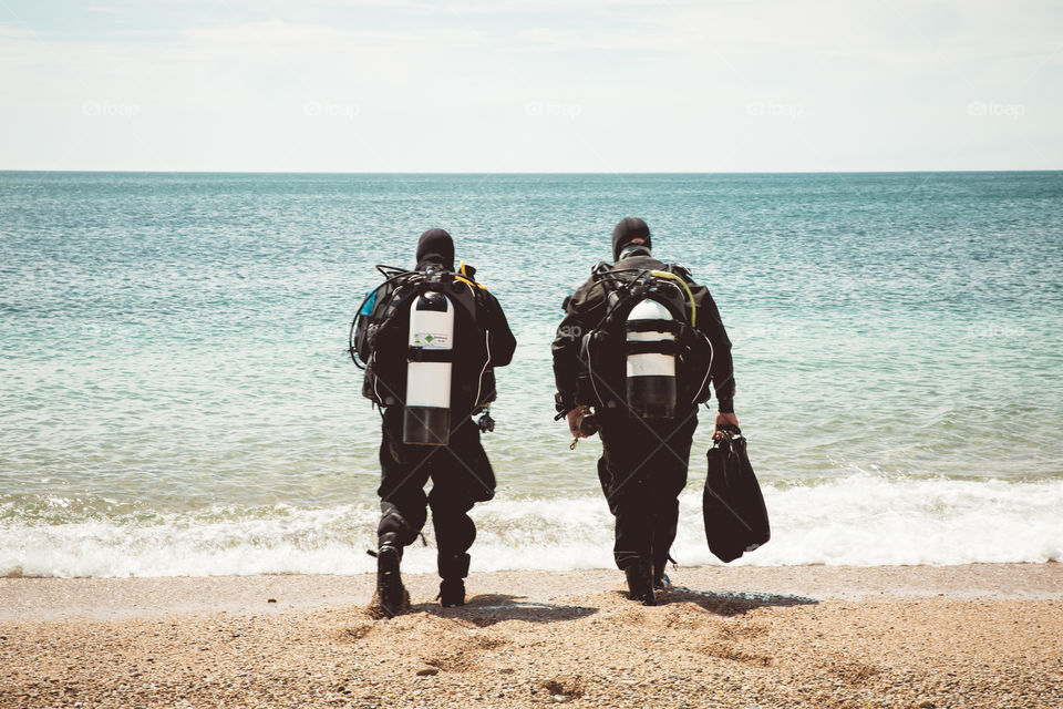 A rear view of two deep sea divers walking into the ocean from a beach