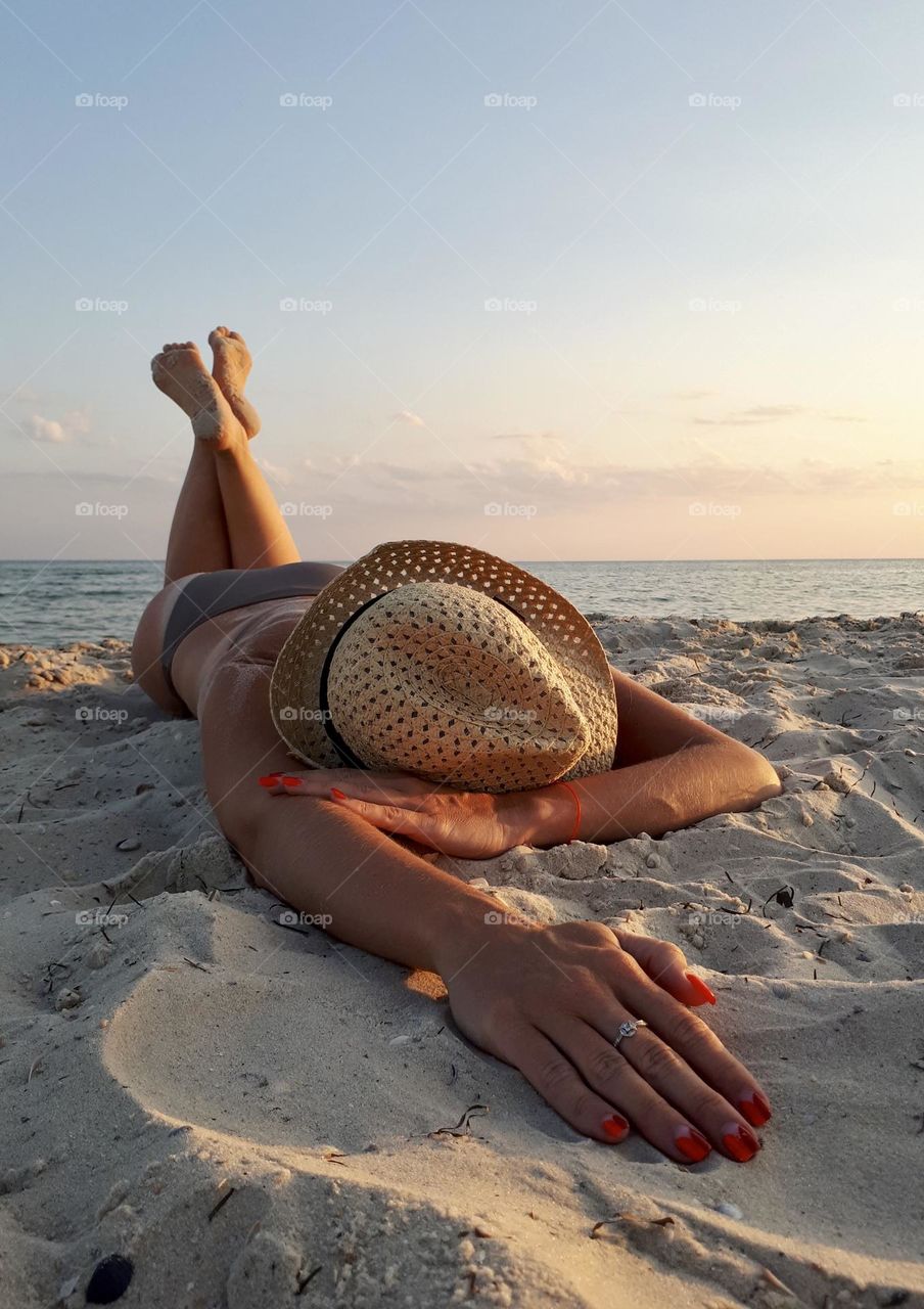 Faceless woman in straw hat and swimming suit lays down on the sunset beach 
