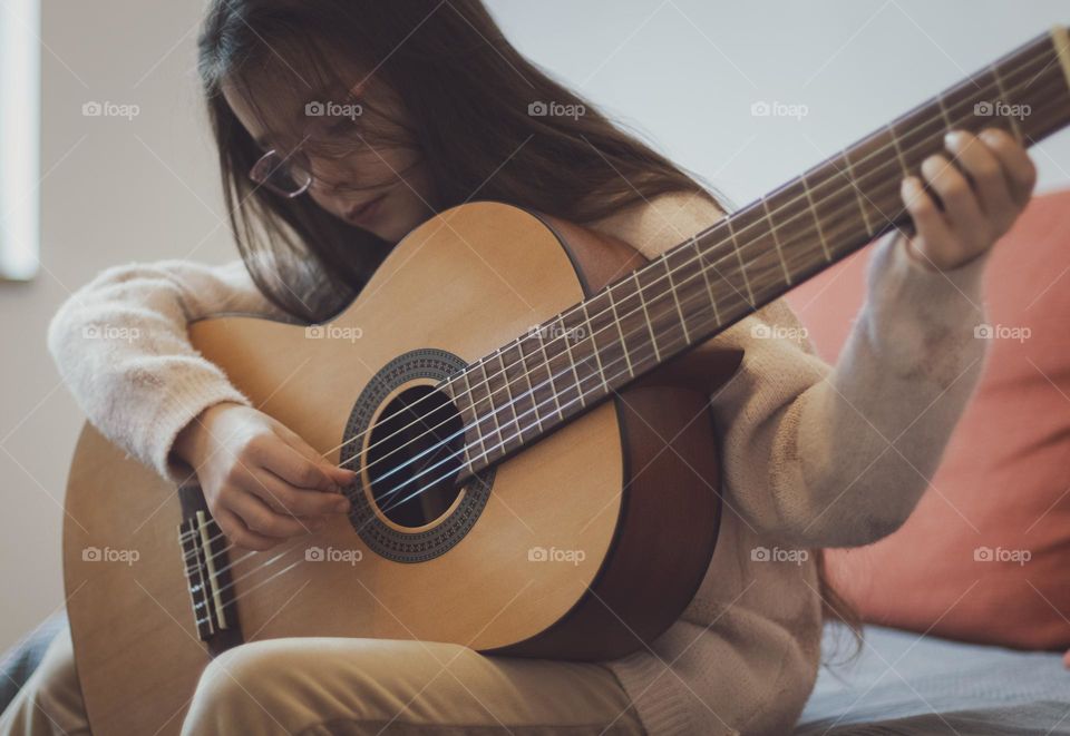 A portrait of one beautiful Caucasian brunette girl with flowing long hair and glasses sits on a sofa in the living room and holds a guitar in an inclination, looking at the strings, sorting through them with her fingers and learning notes, close-up