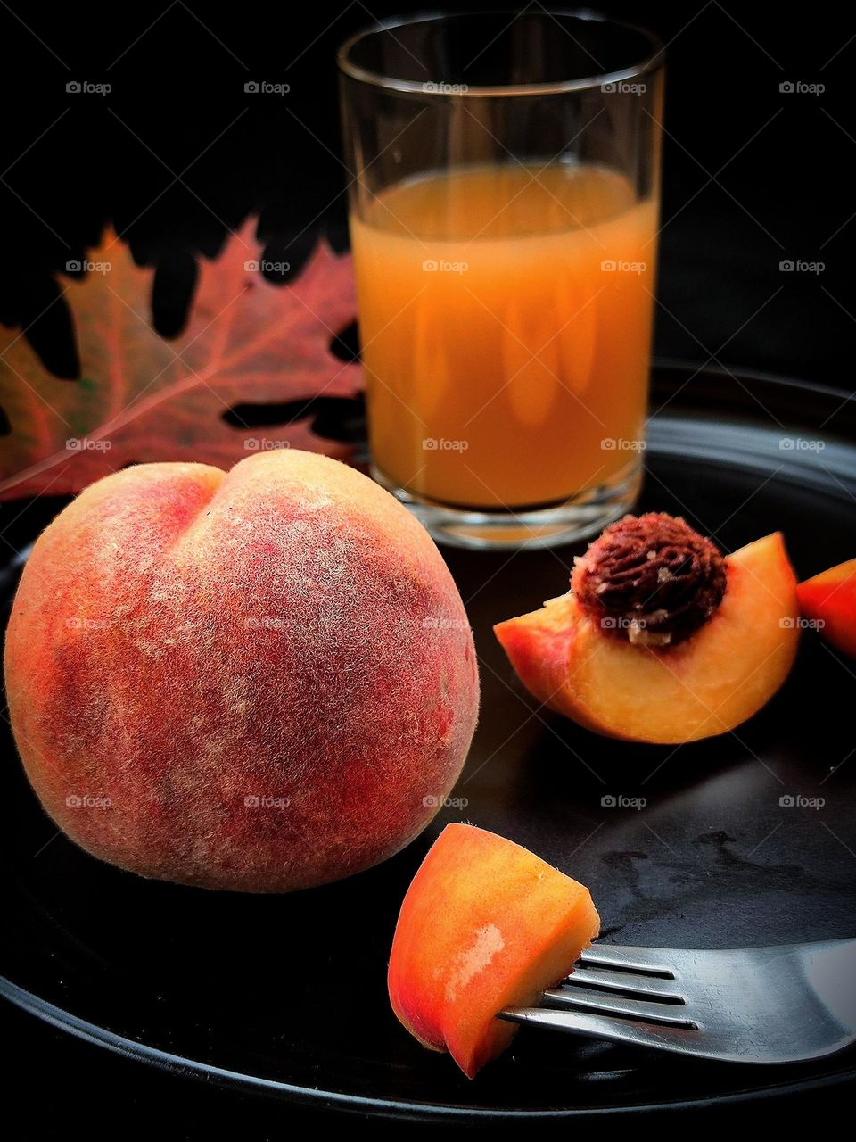 Fruit. All shades of autumn in peach. On a black plate lies a ripe peach, half a peach with a pit, and a glass of peach juice. In the foreground is a fork with a piece of juicy peach. In the background is an autumn oak leaf. Black background