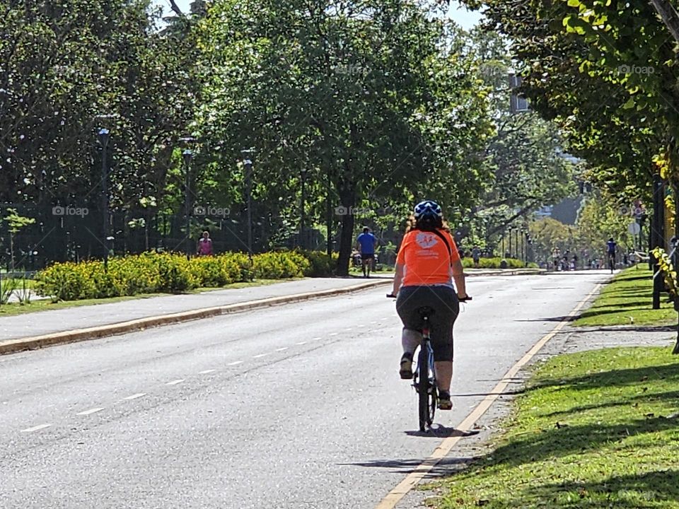 "A lone biker on Sunday". A cyclist with a bright shirt riding on an empty city street, surrounded by grass on one side, and tall trees on the other. A jogger can be seen at the distance. it's a sunny day. A healthy lifestyle.