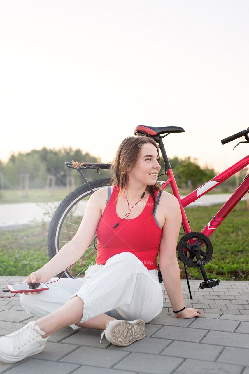 young smiling woman in red shirt and white pants listening to the music standing near her bicycle