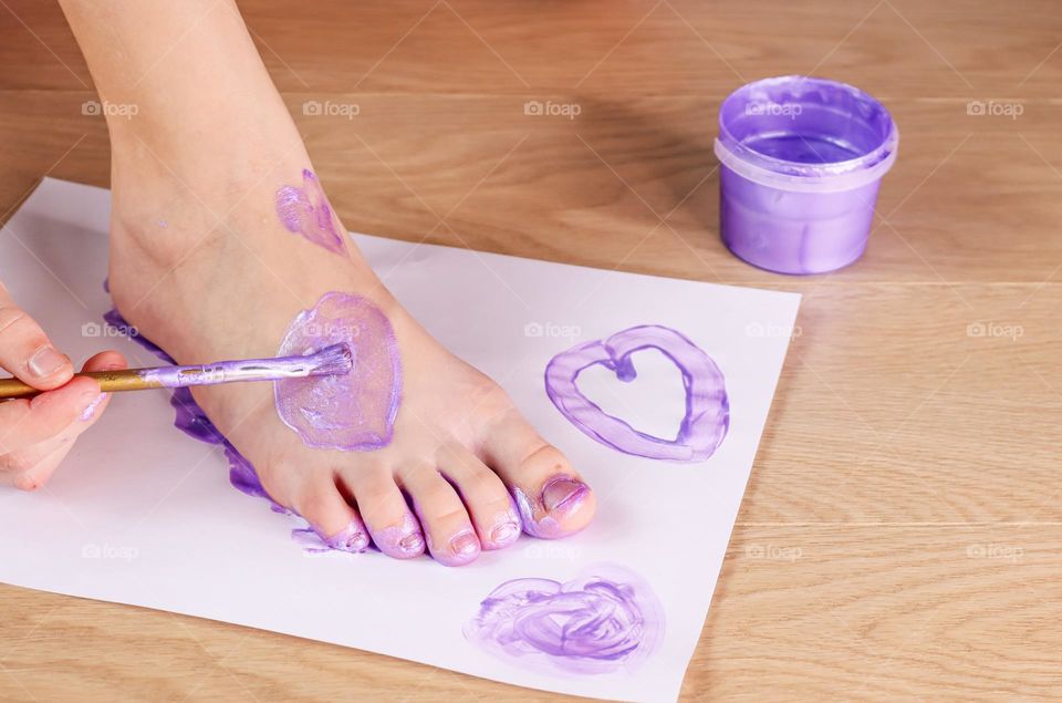 A caucasian teenager girl draws a heart with a brush on her foot with a lavender organic paint on a light wooden floor in the room, close-up side view.