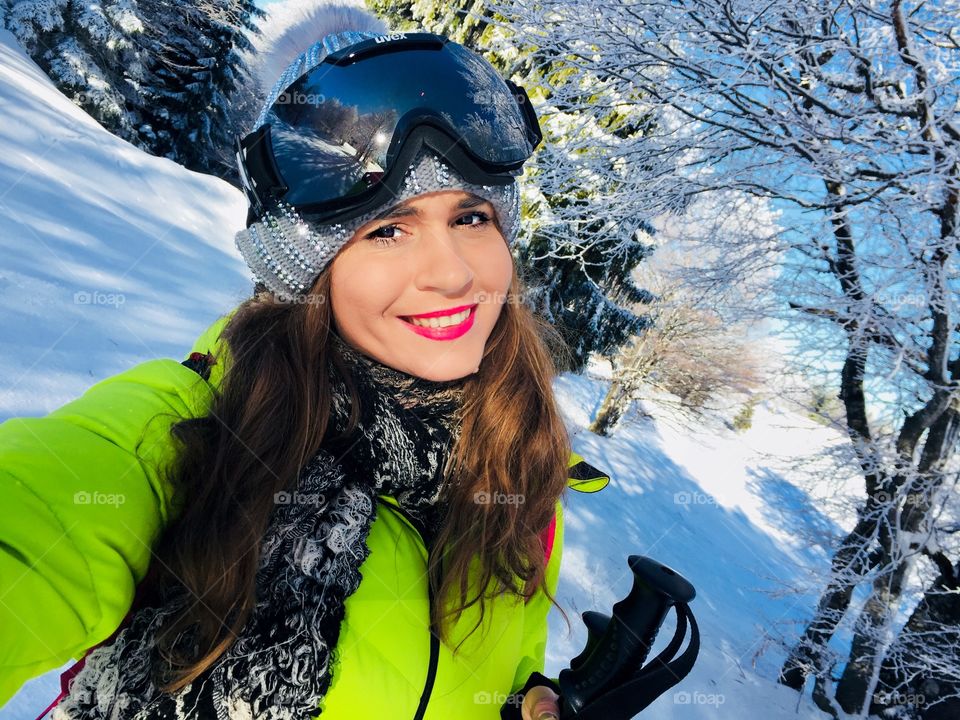 Portrait of smiling young woman wearing Uvex ski glasses with trees covered in snow in the background 
