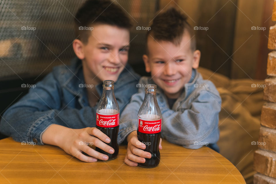 two lovely brothers, dressed in denim clothes, with beautiful hairstyles, are sitting in a cafe and drinking Coca-Cola. communicate, laugh, smile. two guys, two friends