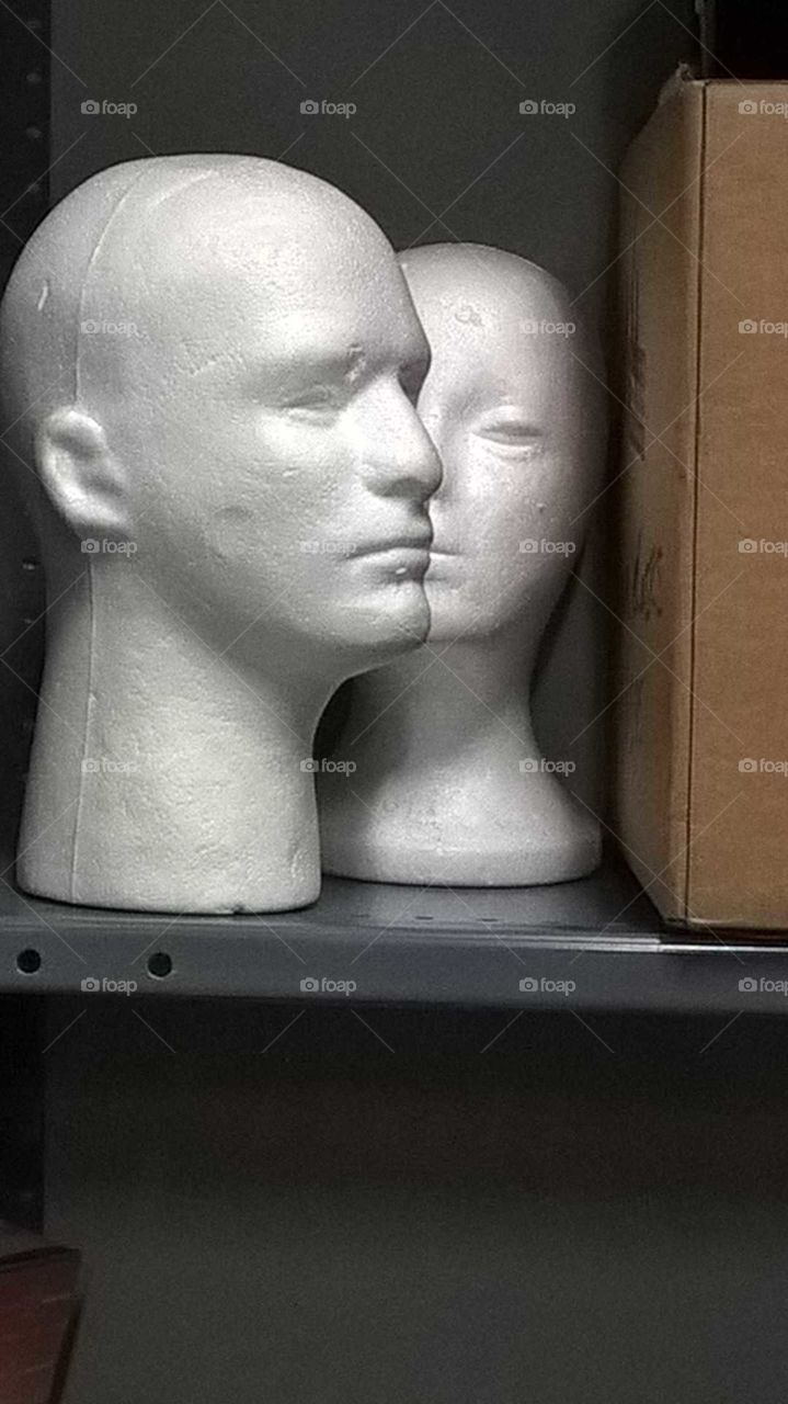 2 styrofoam mannequin heads, whispering together on a stock room shelf.
