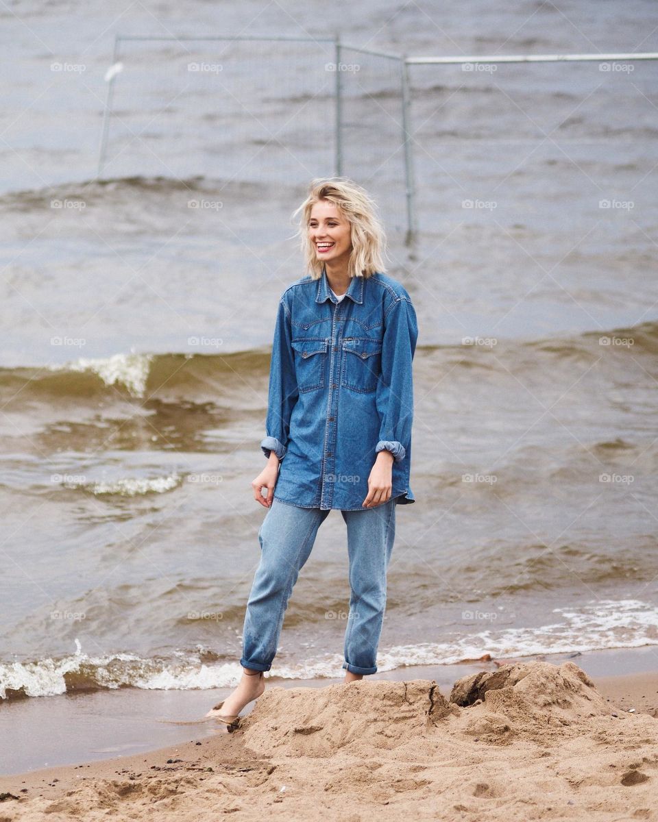Young beautiful smiling girl with blonde hair wearing blue jeans and shirt standing on a sand beach in cloudy summer day, looking in to the distance, portrait of woman 