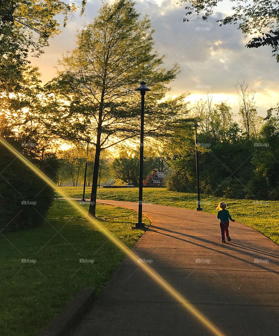 Little Girl running on path at sundown; trees, beams, lamppost 