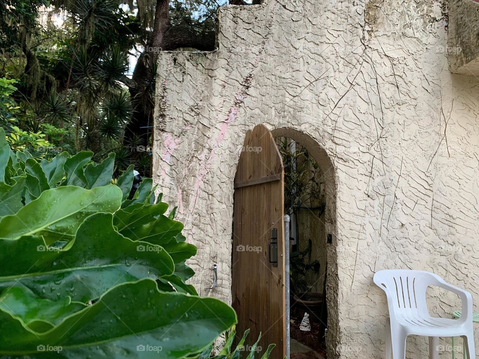 White and red spanish old style architecture residential large house built in the early 1900s. Doorway with antique style wood door in a stairway wall with white chair seen from tropical plants and live oak trees leading to another garden.