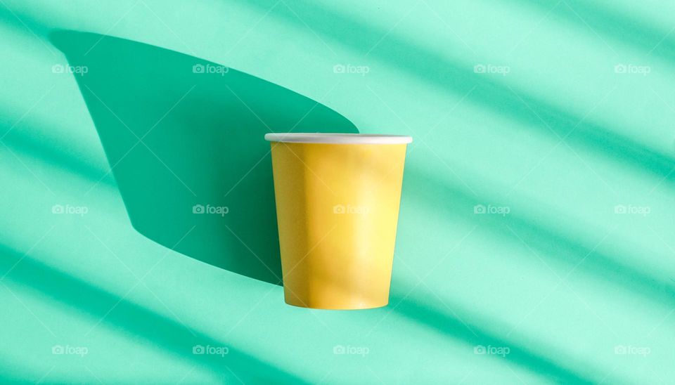 One yellow paper glass lies in the center on a green background with diagonally geometric shadows, flat lay close-up.