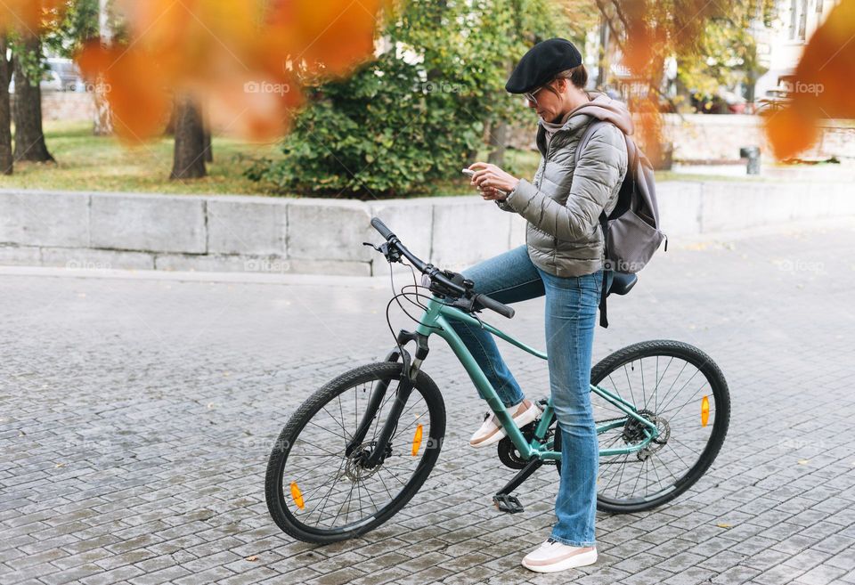 Portrait of fashionable young pretty woman in cap and sunglasses using mobile phone on bicycle on sunny autumn day in city park