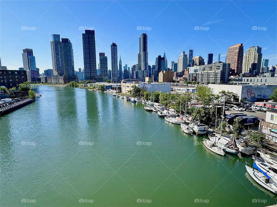 Beautiful “Newtown Creek” in turquoise with a small marina presents itself from the “Pulaski Bridge” that connects “Greenpoint”, Bklyn to LIC, Queens. In the distance we see “Manhattan” incl.  the “Empire State Bldg”. 2024. Hypnotic Productions